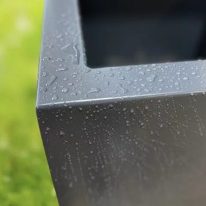 May include: Close-up of a grey metal planter with water droplets on the surface. The planter has a square shape and a smooth, slightly textured finish.