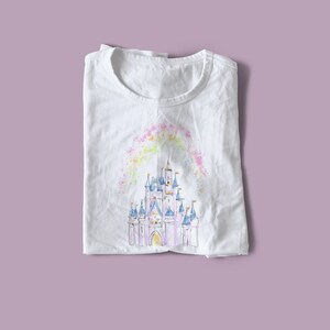 Op de afbeelding: Wit T-shirt met een aquarel-illustratie van een roze kasteel met blauwe torens en een regenboogkleurige lucht erboven.