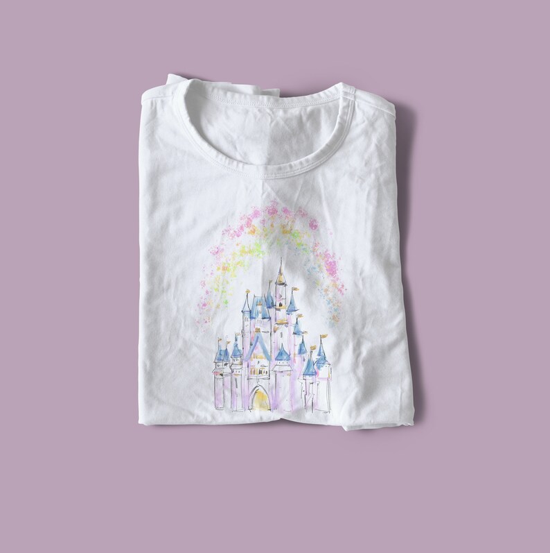 Op de afbeelding: Wit T-shirt met een aquarel-illustratie van een roze kasteel met blauwe torens en een regenboogkleurige lucht erboven.