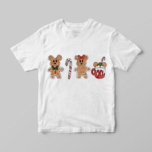 May include: White t-shirt featuring a festive design with gingerbread bear characters, a candy cane, and a mug of hot cocoa. The gingerbread bears have green bow ties and red bows. The hot cocoa mug is red with white whipped cream and a candy cane.