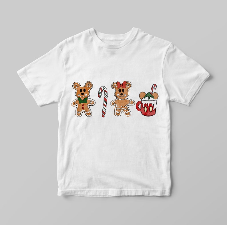 May include: White t-shirt featuring a festive design with gingerbread bear characters, a candy cane, and a mug of hot cocoa. The gingerbread bears have green bow ties and red bows. The hot cocoa mug is red with white whipped cream and a candy cane.