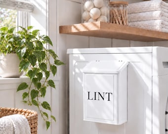May include: A white, rectangular, wall-mounted metal bin with a hinged lid, featuring the word LINT printed in black capital letters on the front. The container is attached to the side of a white appliance, designed for collecting laundry lint.