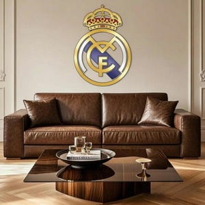 Può includere: Un stemma dorato del Real Madrid con una corona è montato su una parete beige sopra un divano in pelle marrone. Lo stemma presenta un contorno dorato, dettagli bianchi e blu e le lettere "RMCF". Un tavolino in legno scuro è di fronte al divano.
