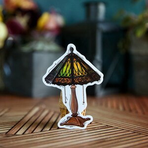 Pu&ograve; includere: Un adesivo di una lampada in vetro colorato con un design verde, giallo e marrone. La lampada ha una base marrone e un cavo nero.