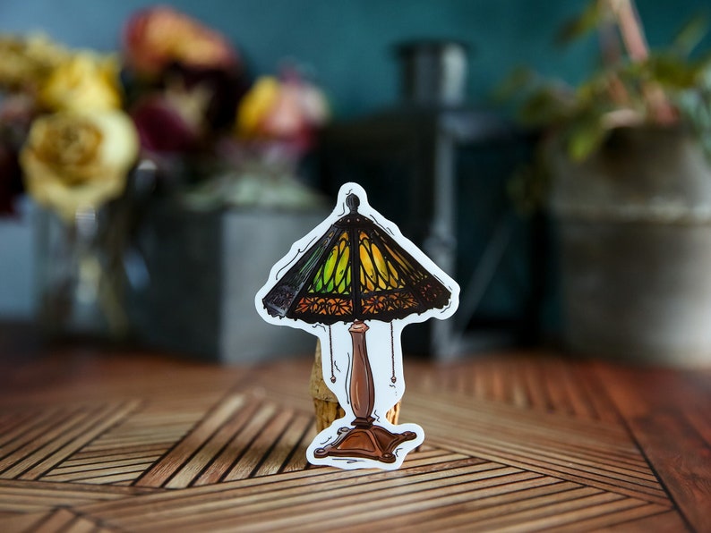 Pu&ograve; includere: Un adesivo di una lampada in vetro colorato con un design verde, giallo e marrone. La lampada ha una base marrone e un cavo nero.