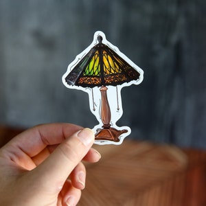 Pu&ograve; includere: Un adesivo di una lampada in vetro colorato con una base marrone e un paralume in vetro colorato giallo, verde e nero.