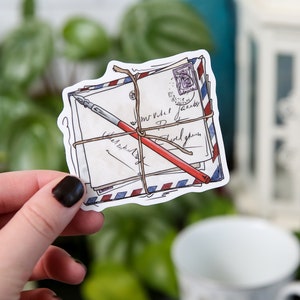 May include: A sticker of a stack of envelopes tied with twine and a red pen. The envelopes have handwritten text and a blue and red stamp.