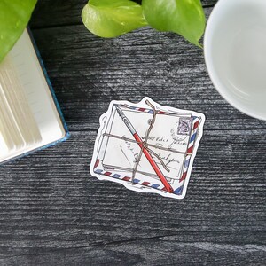 May include: A sticker of a stack of envelopes tied with twine and a red pen. The envelopes have blue and red stripes and a stamp with a picture of a person on it.