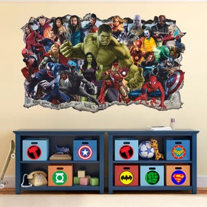 Könnte beinhalten: Ein Wandtattoo mit einer Collage von Superhelden, darunter Hulk, Iron Man und Spider-Man, die aus einer gefälschten Wand herausbrechen. Darunter ein blaues Regal mit Aufbewahrungsboxen mit Superhelden-Logos wie Thors Hammer und Captain Americas Schild.