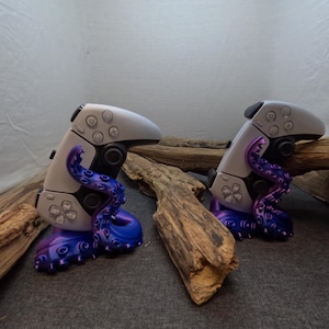 Könnte beinhalten: Zwei weiße Videospielcontroller werden auf einem lila und blauen, krakenförmigen Ständer präsentiert. Die Ständer sind aus 3D-gedrucktem Kunststoff und ruhen auf einem Stück Treibholz.