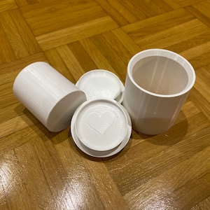 Könnte beinhalten: Ein Satz weißer, 3D-gedruckter Behälter. Das Set umfasst einen zylindrischen Sockel, zwei Deckel und ein separates zylindrisches Teil. Ein Deckel hat ein Herzdesign. Auf einer Holzoberfläche angeordnet.