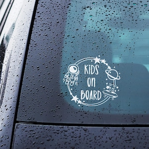 May include: White vinyl car decal with the text "KIDS ON BOARD" inside a circular design. The design includes an astronaut, stars, a planet, and a UFO with an alien. The decal is applied to a car window.