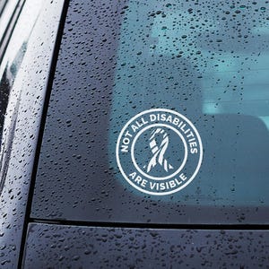 Op de afbeelding: Een witte, ronde sticker op een autoruit met de tekst "NOT ALL DISABILITIES ARE VISIBLE" in een cirkelvormig ontwerp. De sticker heeft een lintafbeelding in het midden en is bevestigd op een zwarte auto.