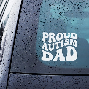 May include: White vinyl decal with the words "PROUD AUTISM DAD" in a retro font, affixed to a car window. The window is covered in water droplets, and the car is dark blue.