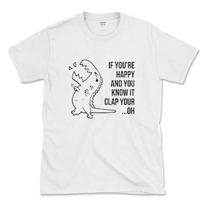 Peut inclure: T-shirt blanc avec une illustration noir et blanc d'un dinosaure en train de pleurer. Le texte "IF YOU'RE HAPPY AND YOU KNOW IT CLAP YOUR ...OH" est imprim&eacute; sur le t-shirt.