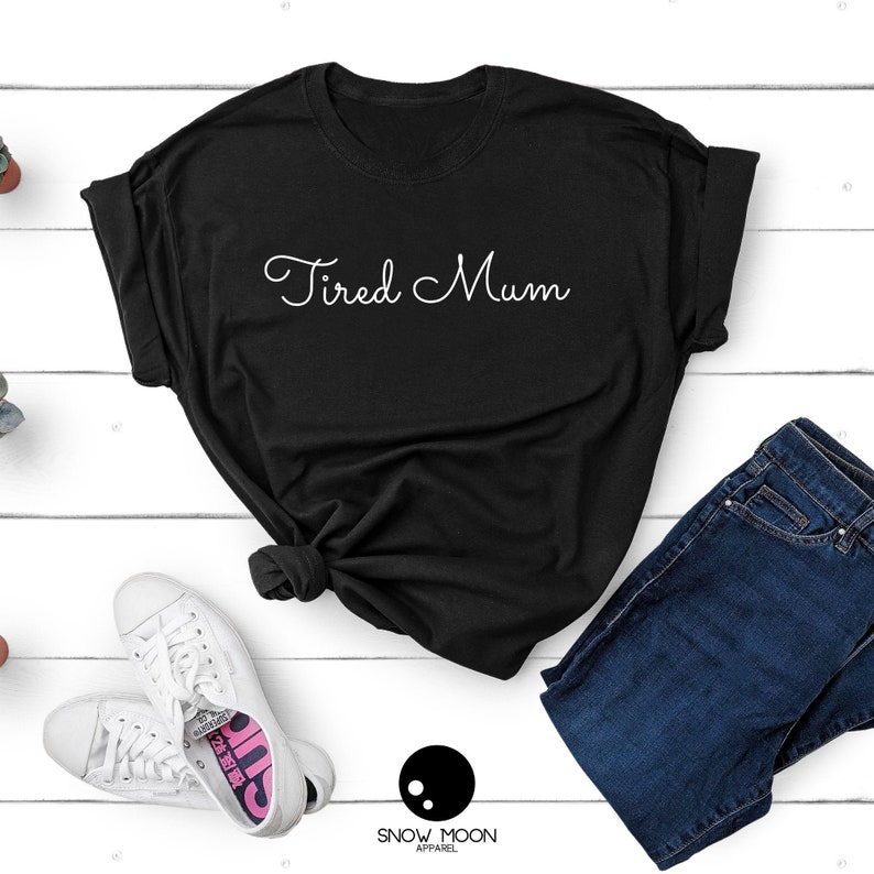 May include: Black t-shirt with the text "Tired Mum" in white cursive writing. The shirt is laid flat on a white wooden surface with a pair of white sneakers, blue jeans, and a small plant.