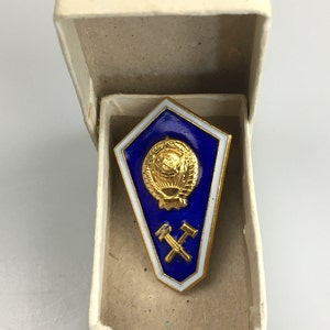 Puede incluir: Un pin de solapa en forma de escudo con un fondo de esmalte azul intenso y un borde blanco. El pin muestra un emblema dorado de un globo terráqueo rodeado de espigas de trigo sobre un par de martillos cruzados, dentro de una caja color crema.