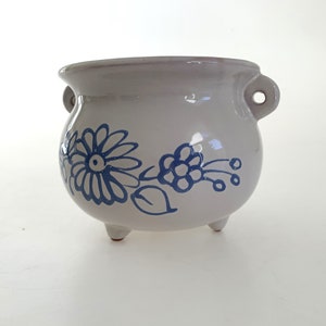 May include: White ceramic pot with blue floral design. The pot has three legs and two handles.