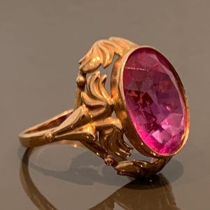 K&ouml;nnte beinhalten: Ein goldfarbener Ring mit einem ovalen, facettierten, rosafarbenen Edelstein. Der Ring hat ein verziertes, blumeninspiriertes Design um den Edelstein und am Band. Der Ring wird auf einer reflektierenden Oberfl&auml;che pr&auml;sentiert.