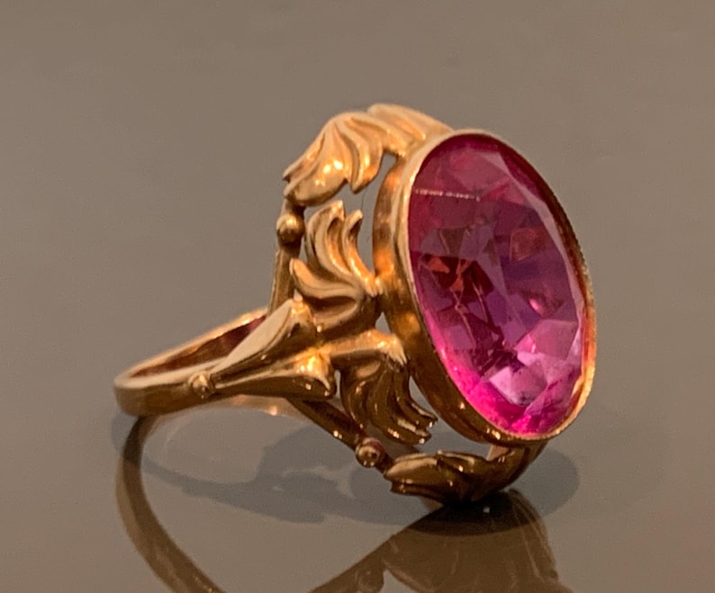 K&ouml;nnte beinhalten: Ein goldfarbener Ring mit einem ovalen, facettierten, rosafarbenen Edelstein. Der Ring hat ein verziertes, blumeninspiriertes Design um den Edelstein und am Band. Der Ring wird auf einer reflektierenden Oberfl&auml;che pr&auml;sentiert.