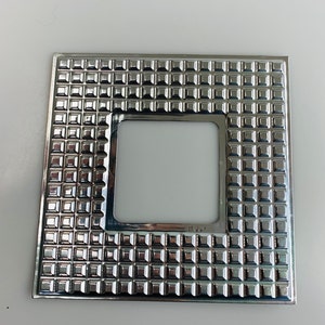 Puede incluir: Un marco cuadrado plateado con un patrón texturizado en relieve de pequeños cuadrados. El marco tiene una abertura cuadrada en el centro.