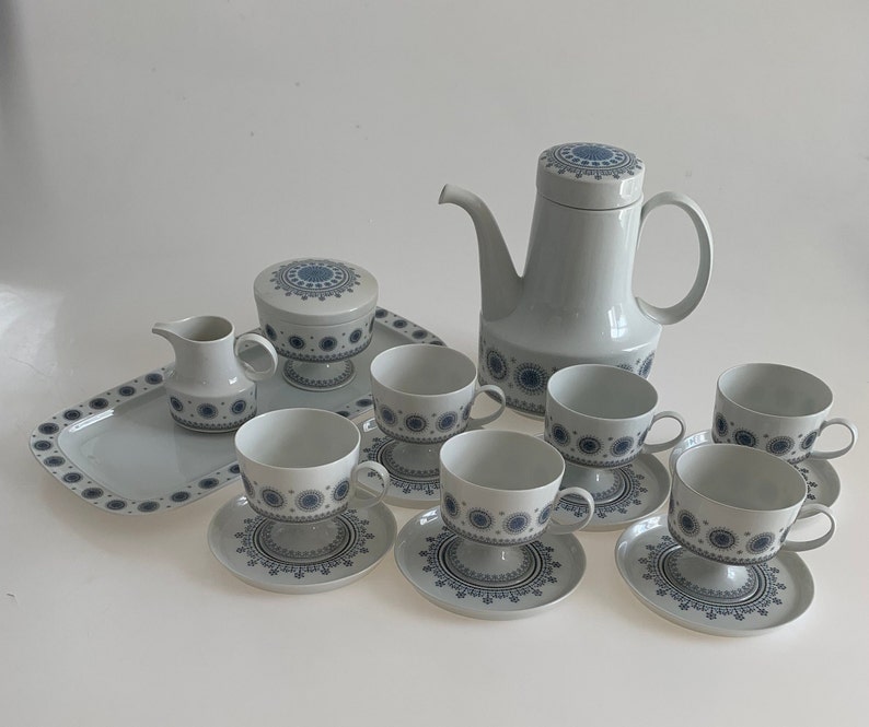 K&ouml;nnte beinhalten: Ein wei&szlig;es Teeservice mit blauen Blumendesigns. Das Set beinhaltet eine Teekanne, eine Zuckerdose, eine Milchk&auml;nnchen und sechs Tassen und Untertassen.