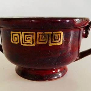 Pode incluir: Uma tigela de sopa de cerâmica vermelho escuro com duas alças e um design geométrico dourado.