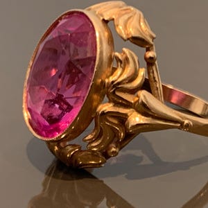 K&ouml;nnte beinhalten: Ein goldfarbener Ring mit einem gro&szlig;en, oval geschliffenen, rosafarbenen Edelstein. Der Ring hat verzierte, wirbelnde Details um den Edelstein und am Band. Der Ring ist wahrscheinlich Vintage oder antik.