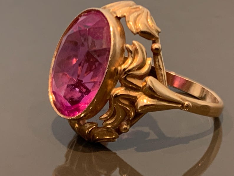 K&ouml;nnte beinhalten: Ein goldfarbener Ring mit einem gro&szlig;en, oval geschliffenen, rosafarbenen Edelstein. Der Ring hat verzierte, wirbelnde Details um den Edelstein und am Band. Der Ring ist wahrscheinlich Vintage oder antik.