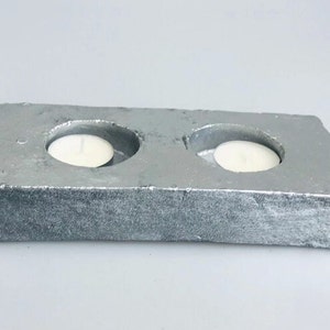 Puede incluir: Un portavelas de hormigón plateado con dos depresiones circulares para velas votivas.