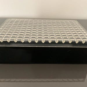 Può includere: Una scatola rettangolare nera con un coperchio in plastica trasparente. Il coperchio ha un motivo a griglia.