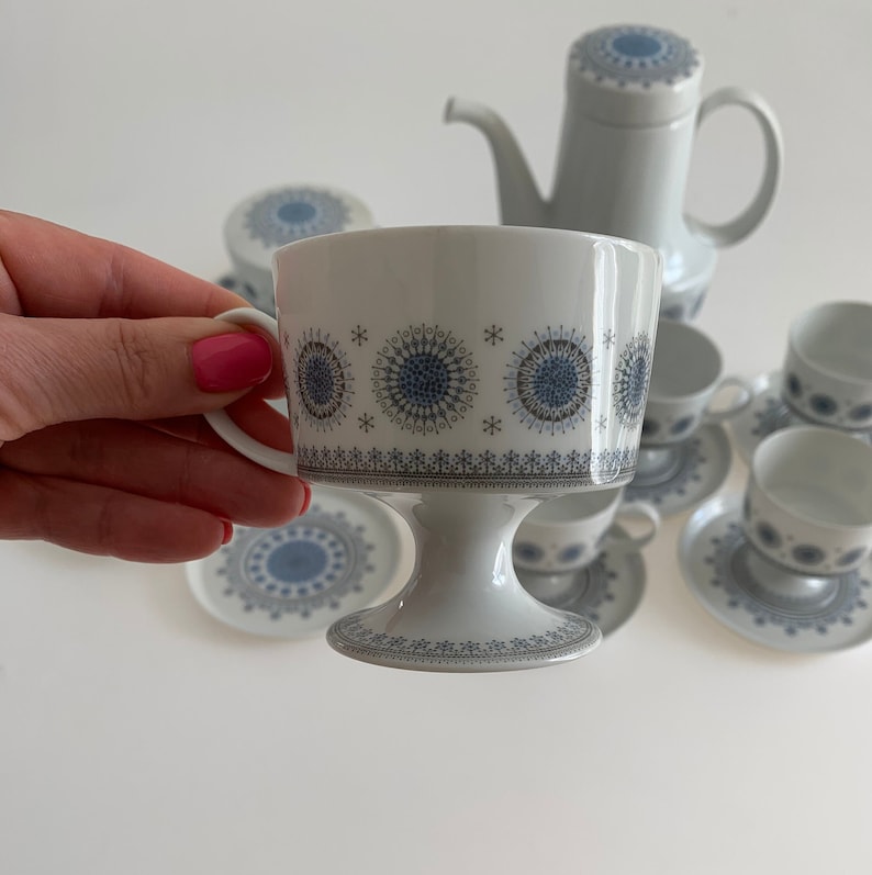 K&ouml;nnte beinhalten: Eine wei&szlig;e Teetasse und Untertasse mit einem blauen und grauen geometrischen Muster. Die Tasse hat einen Sockel und die Untertasse einen gewellten Rand.