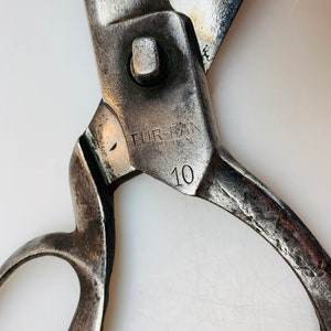 May include: A pair of heavy-duty metal shears with a weathered, dark silver finish. The pivot area features the stamped text TUR-BAN and the number 10. The blades are partially open, showing signs of wear and oxidation on the metal surface.