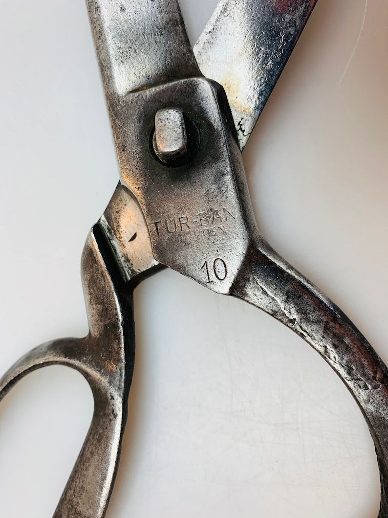 May include: A pair of heavy-duty metal shears with a weathered, dark silver finish. The pivot area features the stamped text TUR-BAN and the number 10. The blades are partially open, showing signs of wear and oxidation on the metal surface.