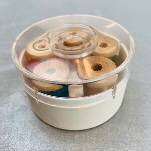 May include: A round, white plastic container with a clear, removable lid. Inside the container are several small, wooden spools of thread in various colors, including blue and red, arranged in a circular pattern within the storage unit.