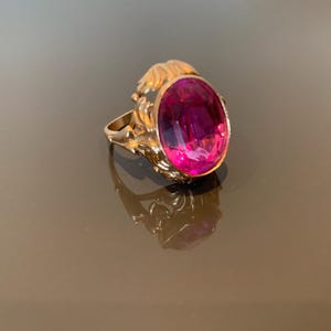 K&ouml;nnte beinhalten: Ein goldener Ring mit einem gro&szlig;en, ovalen, magentafarbenen Edelstein. Der Ring hat eine verzierte, strukturierte Fassung mit floralem Design. Der Ring wird auf einer reflektierenden Oberfl&auml;che pr&auml;sentiert.