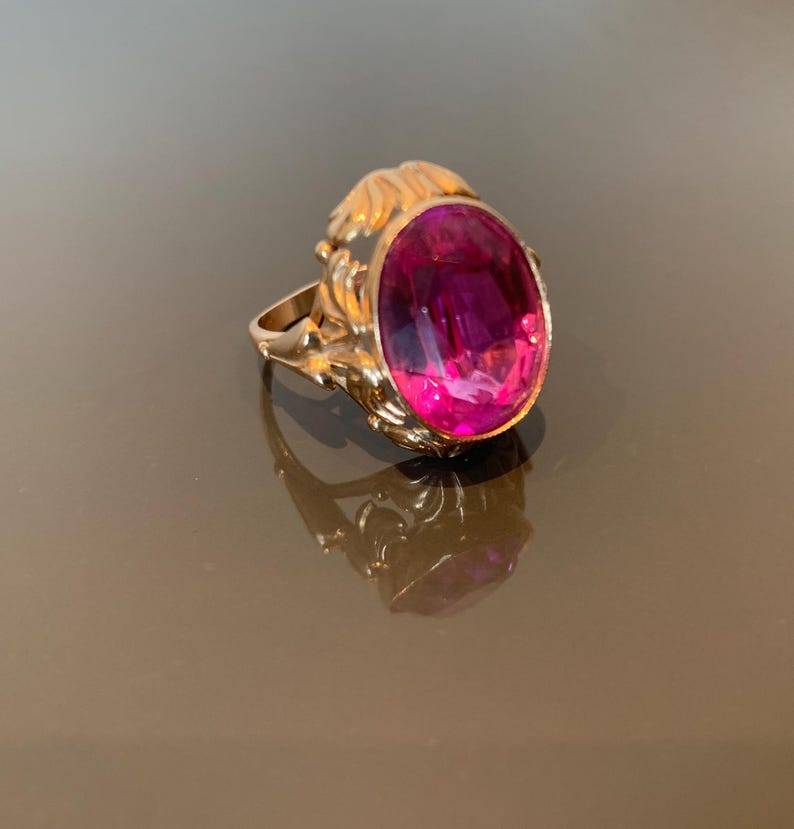K&ouml;nnte beinhalten: Ein goldener Ring mit einem gro&szlig;en, ovalen, magentafarbenen Edelstein. Der Ring hat eine verzierte, strukturierte Fassung mit floralem Design. Der Ring wird auf einer reflektierenden Oberfl&auml;che pr&auml;sentiert.