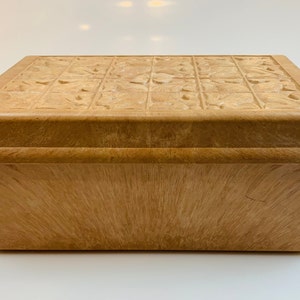 Könnte beinhalten: Eine Holzkiste mit einem geschnitzten Blumendesign auf dem Deckel. Die Kiste ist hellbraun und hat eine glatte Oberfläche.