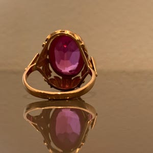 K&ouml;nnte beinhalten: Ein goldener Ring mit einem gro&szlig;en, oval geschliffenen, tiefrosa Edelstein. Der Ring hat eine verzierte Fassung mit blattartigen Akzenten an den Schultern. Der Edelstein reflektiert das Licht und zeigt seine Facetten. Der Ring wird auf einer reflektierenden Oberfl&auml;che gezeigt.