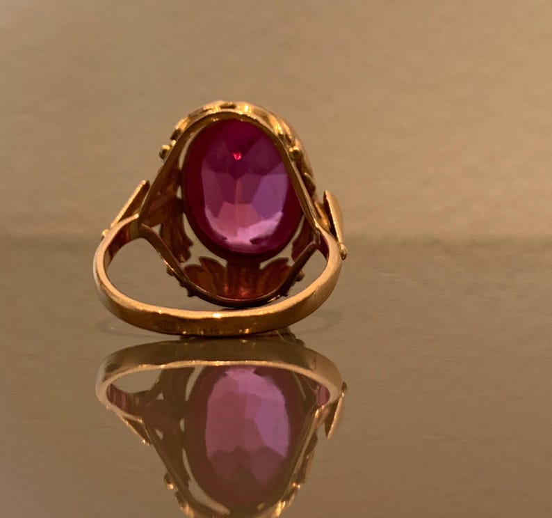 K&ouml;nnte beinhalten: Ein goldener Ring mit einem gro&szlig;en, oval geschliffenen, tiefrosa Edelstein. Der Ring hat eine verzierte Fassung mit blattartigen Akzenten an den Schultern. Der Edelstein reflektiert das Licht und zeigt seine Facetten. Der Ring wird auf einer reflektierenden Oberfl&auml;che gezeigt.