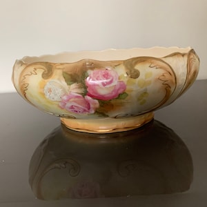 May include: A porcelain bowl featuring a scalloped rim and a hand-painted design of pink roses with green leaves. The exterior has a cream-coloured base with gold-toned scrollwork accents and a gold-trimmed pedestal base, creating a decorative piece.