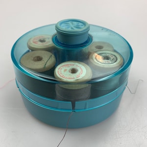 Puede incluir: Un contenedor redondo de plástico azul translúcido para almacenar hilo de coser, con una tapa de pomo central. En su interior se ven varios carretes de hilo de madera, cada uno con una etiqueta de papel circular con texto y un orificio.