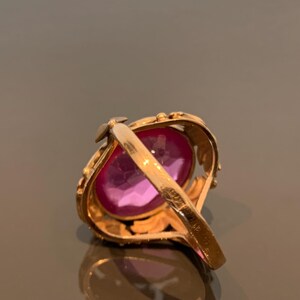 K&ouml;nnte beinhalten: Ein goldfarbener Ring mit einem gro&szlig;en, oval geschliffenen, rosafarbenen Edelstein. Das Band des Rings hat ein verziertes, blattartiges Design. Der Edelstein ist der Mittelpunkt und reflektiert einen leuchtenden rosa Farbton. Der Ring ist vor einem neutralen Hintergrund.