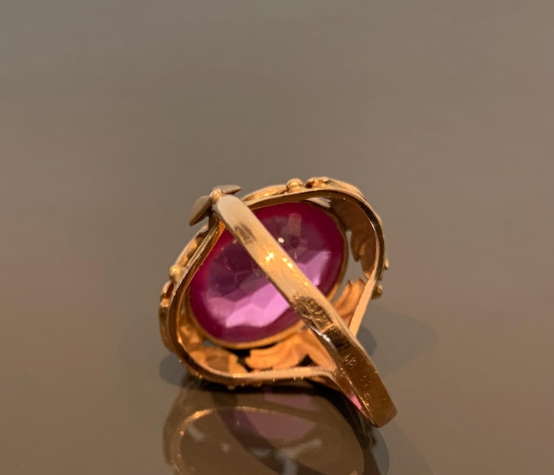 K&ouml;nnte beinhalten: Ein goldfarbener Ring mit einem gro&szlig;en, oval geschliffenen, rosafarbenen Edelstein. Das Band des Rings hat ein verziertes, blattartiges Design. Der Edelstein ist der Mittelpunkt und reflektiert einen leuchtenden rosa Farbton. Der Ring ist vor einem neutralen Hintergrund.