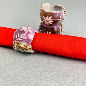 Op de afbeelding: Twee servetringen van schelpen in paars en bruin met een bloemenmotief. De ringen liggen op een rode servet.
