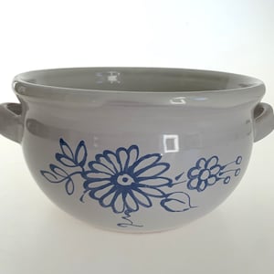 Op de afbeelding: Witte keramische kom met een blauw bloemenontwerp. De kom heeft twee handvatten en een afgeronde vorm.