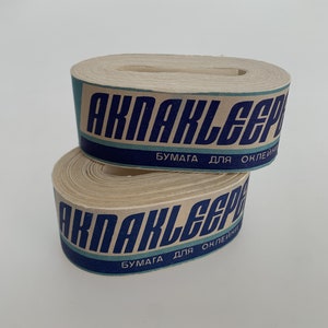 Puede incluir: Dos rollos de cinta adhesiva de papel marrón con letras azules que dicen "AKPAKLEEP" y "БУМАГА ДЛЯ ОКЛЕИНИ" (Papel para pegar).