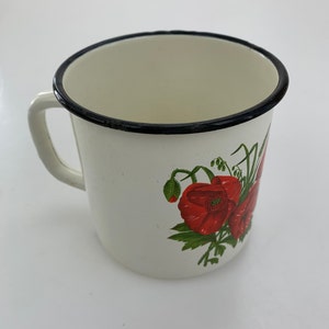 Puede incluir: Una taza de esmalte blanco con un borde negro y un asa lateral. El exterior está decorado con una ilustración botánica de vibrantes flores de amapola rojas con tallos y hojas verdes, mostrando un diseño clásico sobre una superficie lisa.