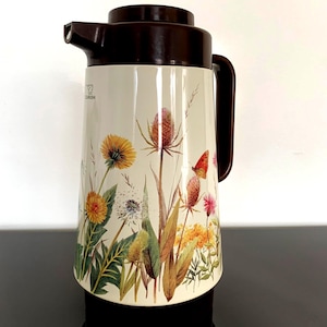 Puede incluir: Un termo vintage de color crema con tapa, asa y boquilla marrones. El termo está decorado con un diseño floral con flores silvestres amarillas, naranjas y rosas, hojas verdes y una mariposa. Está diseñado para contener bebidas frías o calientes.