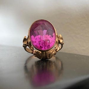 K&ouml;nnte beinhalten: Ein goldfarbener Ring mit einem gro&szlig;en, oval geschliffenen, facettierten rosa Edelstein. Der Ring hat ein verziertes, florales Design am Band und der Fassung. Der Edelstein reflektiert das Licht und erzeugt einen funkelnden Effekt.